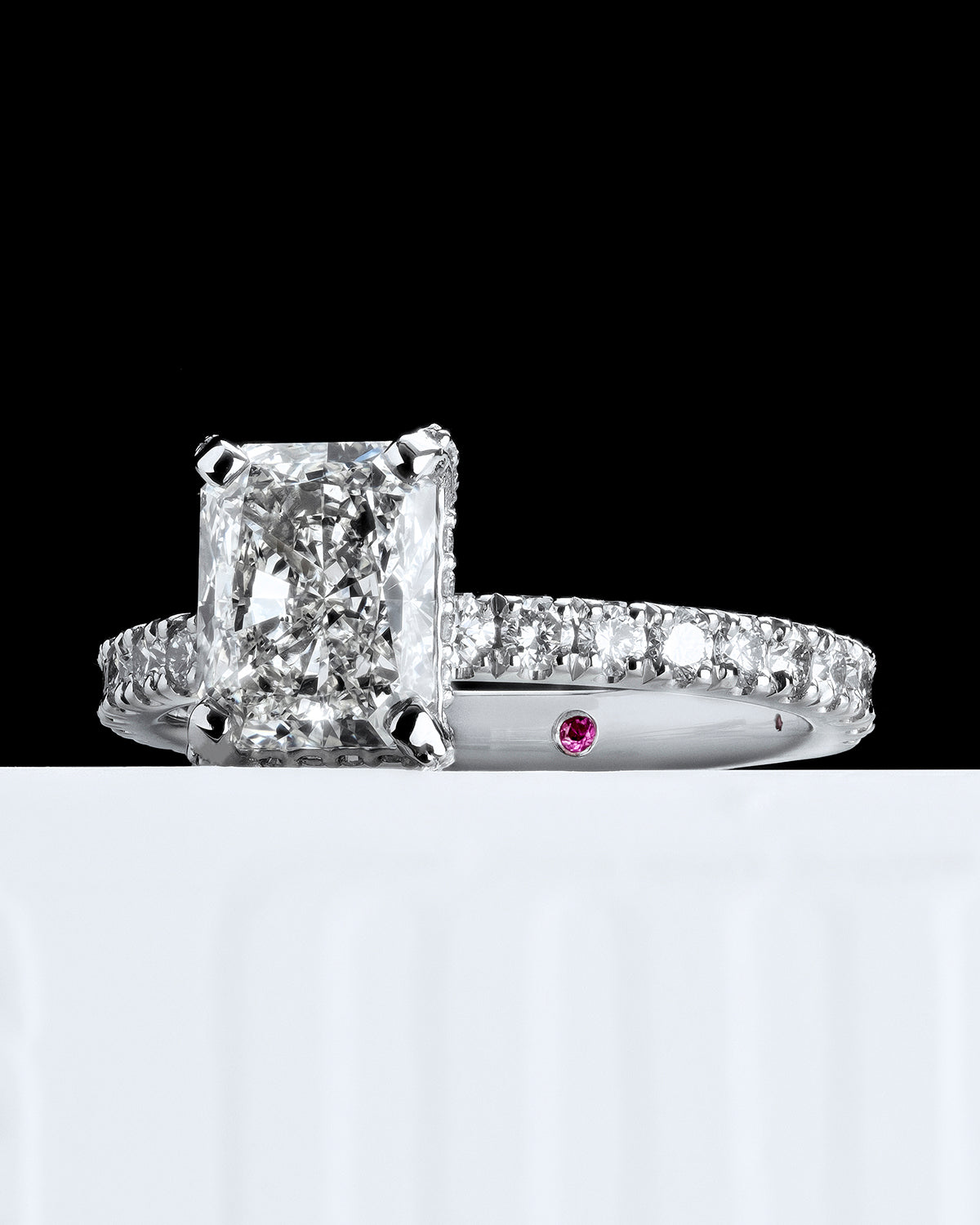 Close-up of a radiant-cut diamond engagement ring featuring a pavé diamond band in polished white gold. The centre stone's sharp facets are beautifully complemented by the intricate pavé detailing. A hidden pink diamond is set within the band, symbolising a unique and personal touch. The composition showcases the brilliance of the diamonds against a sleek, modern background, making this ring ideal for those seeking a luxurious and distinctive engagement ring design.