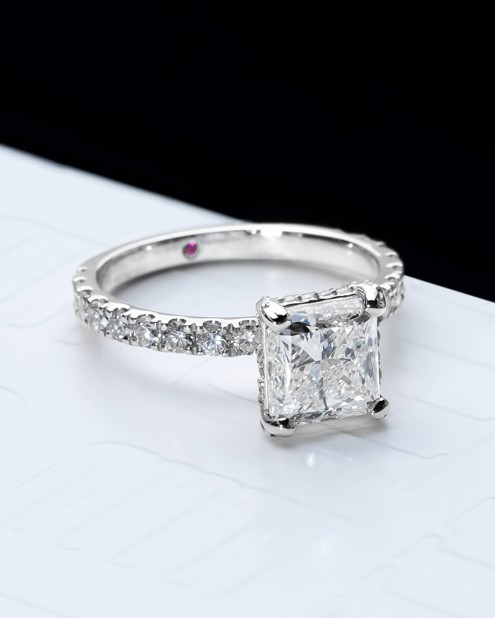 Close-up of a princess-cut diamond engagement ring featuring a pavé diamond band in polished white gold. The square centre stone is elegantly framed by the pavé-set diamonds along the band. A hidden pink diamond inside the band symbolises a rare and personal touch. The ring is displayed on a sleek, embossed MJ Jones surface, highlighting its modern yet timeless appeal. Ideal for those seeking a luxurious and distinctive engagement ring design.