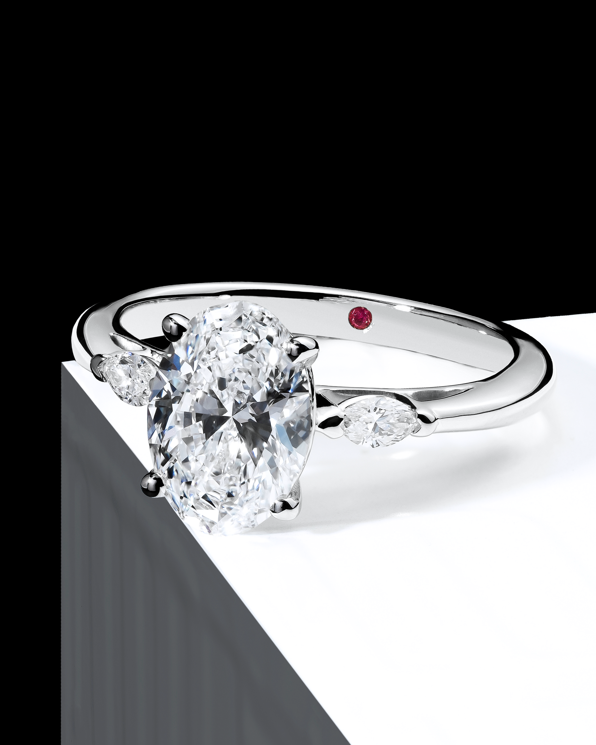 lose-up of an oval-cut diamond engagement ring with a brilliant oval centre stone accompanied by two marquise-shaped diamonds. The polished white gold band features a hidden pink diamond, symbolising a unique touch. The composition highlights the ring's elegant symmetry and sparkle, set against a high-contrast background. Ideal for those seeking an engagement ring that blends classic beauty with contemporary design.
